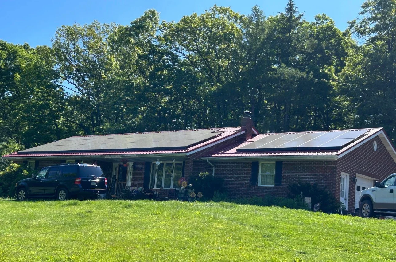 Solar panels installed on brick ranch home with carport and wooded backdrop