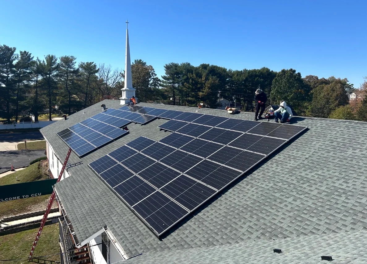Solar panels installed on the flat rooftop of a nonprofit community building in Reading, PA, with rows of panels angled toward the south under a partly cloudy Pennsylvania sky. The building exterior shows institutional construction typical of Berks County, with the solar array covering the majority of the available roof surface.